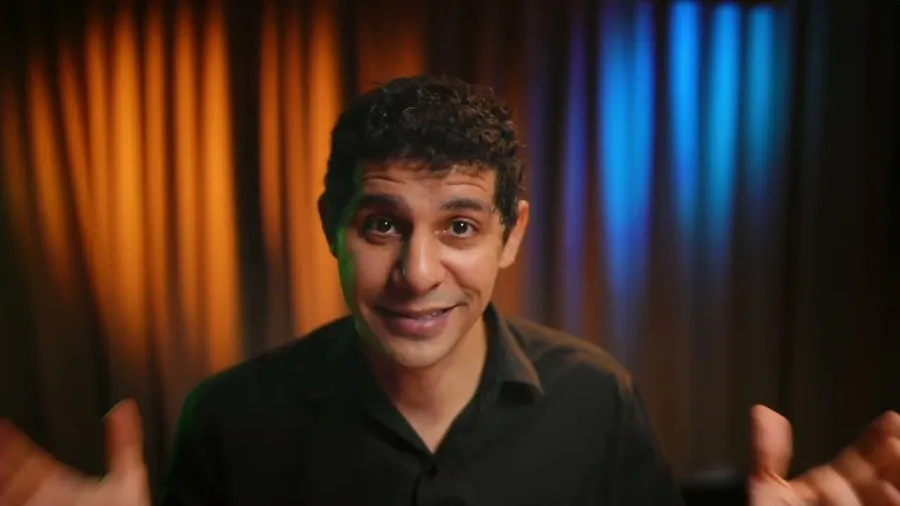 Apresentador da Life Studios falando para a câmera no estúdio, com cortina iluminada em laranja e azul ao fundo.